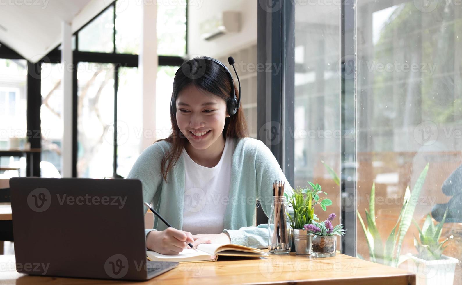 Smiling girl student wear wireless headphone study online with skype teacher, happy young woman learn language listen lecture watch webinar write notes look at laptop sit in cafe, distant education photo