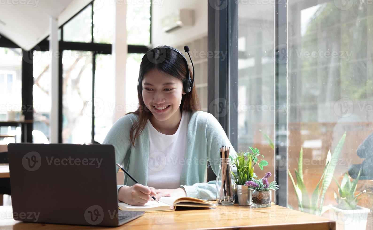 una estudiante sonriente usa auriculares inalámbricos para estudiar en línea con un profesor de skype, una joven feliz aprende el idioma escucha una conferencia mira un seminario web escribe notas mira una laptop sentada en un café, educación distante foto