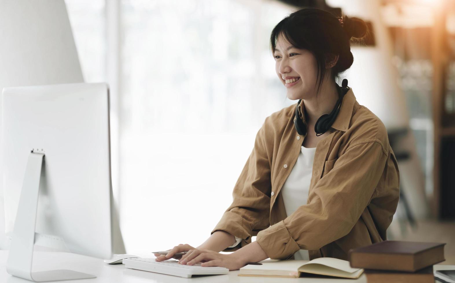 Head shot portrait smiling asian woman wearing headphones posing for photo at workplace, happy excited female wearing headset looking at laptop, sitting at desk with laptop, making video call