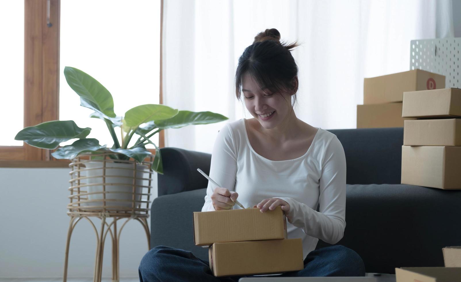 Smiling young Asian business owner woman prepare parcel box and standing check online orders of product for deliver to customer on laptop computer. Shopping Online concept. photo