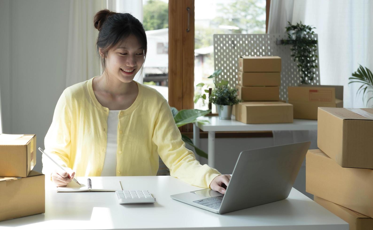 A portrait of Asian woman, e-commerce employee sitting in the office full of packages on the table using a laptop and calculator, for SME business, e-commerce, technology and delivery business. photo