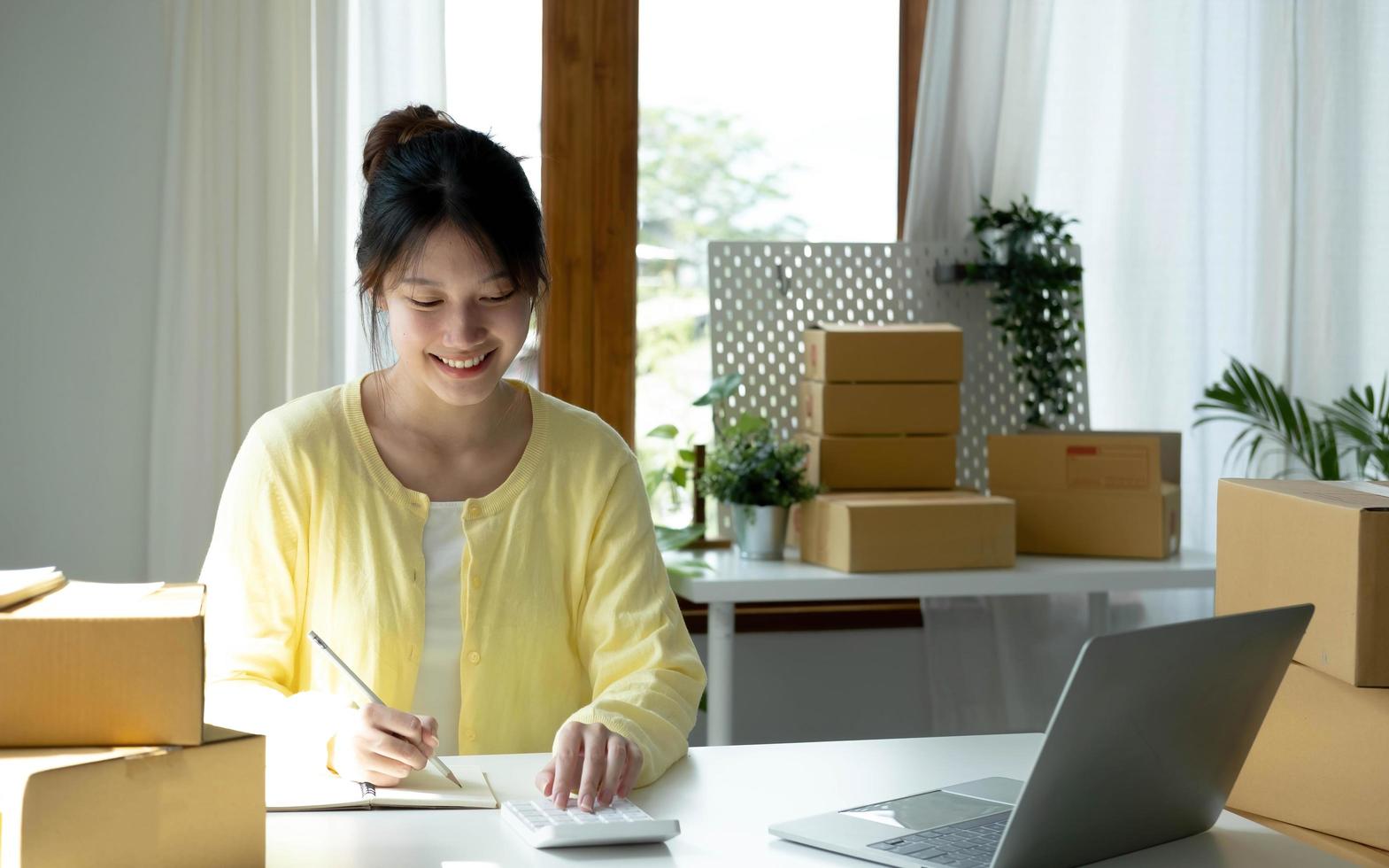 A portrait of Asian woman, e-commerce employee sitting in the office full of packages on the table using a calculator, for SME business, e-commerce, technology and delivery business. photo