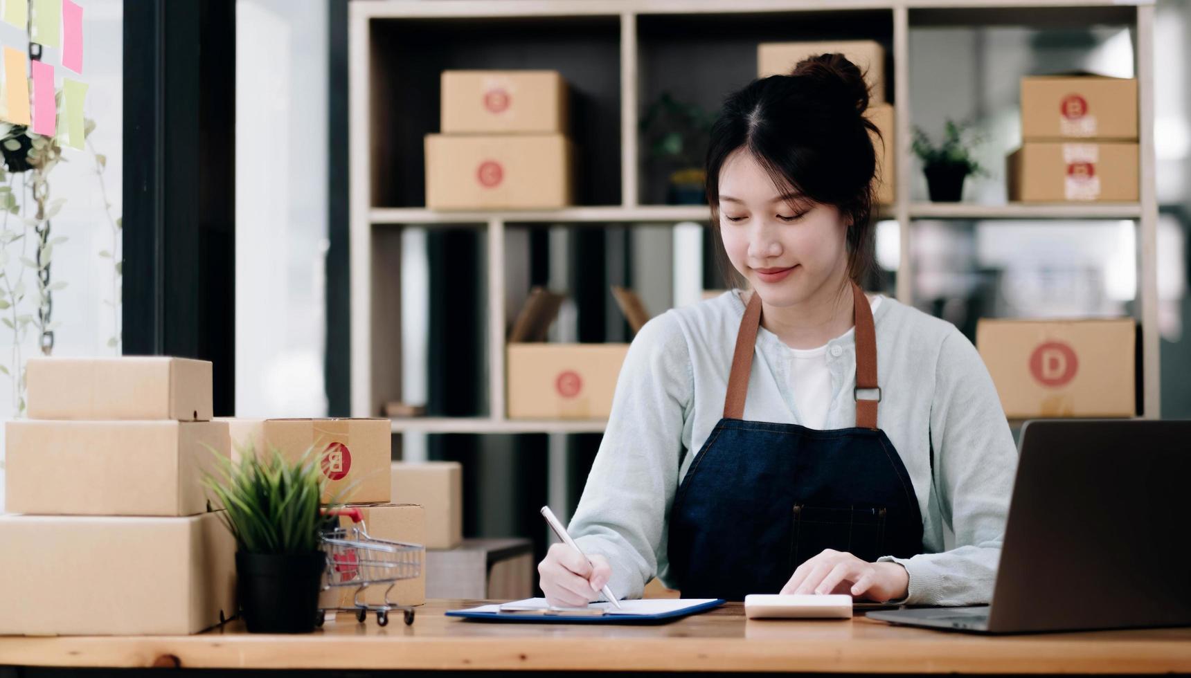 A portrait of a young Asian woman e-commerce employee sitting in the office full of packages in the background write note of orders and a calculator, for SME business e-commerce and delivery business. photo