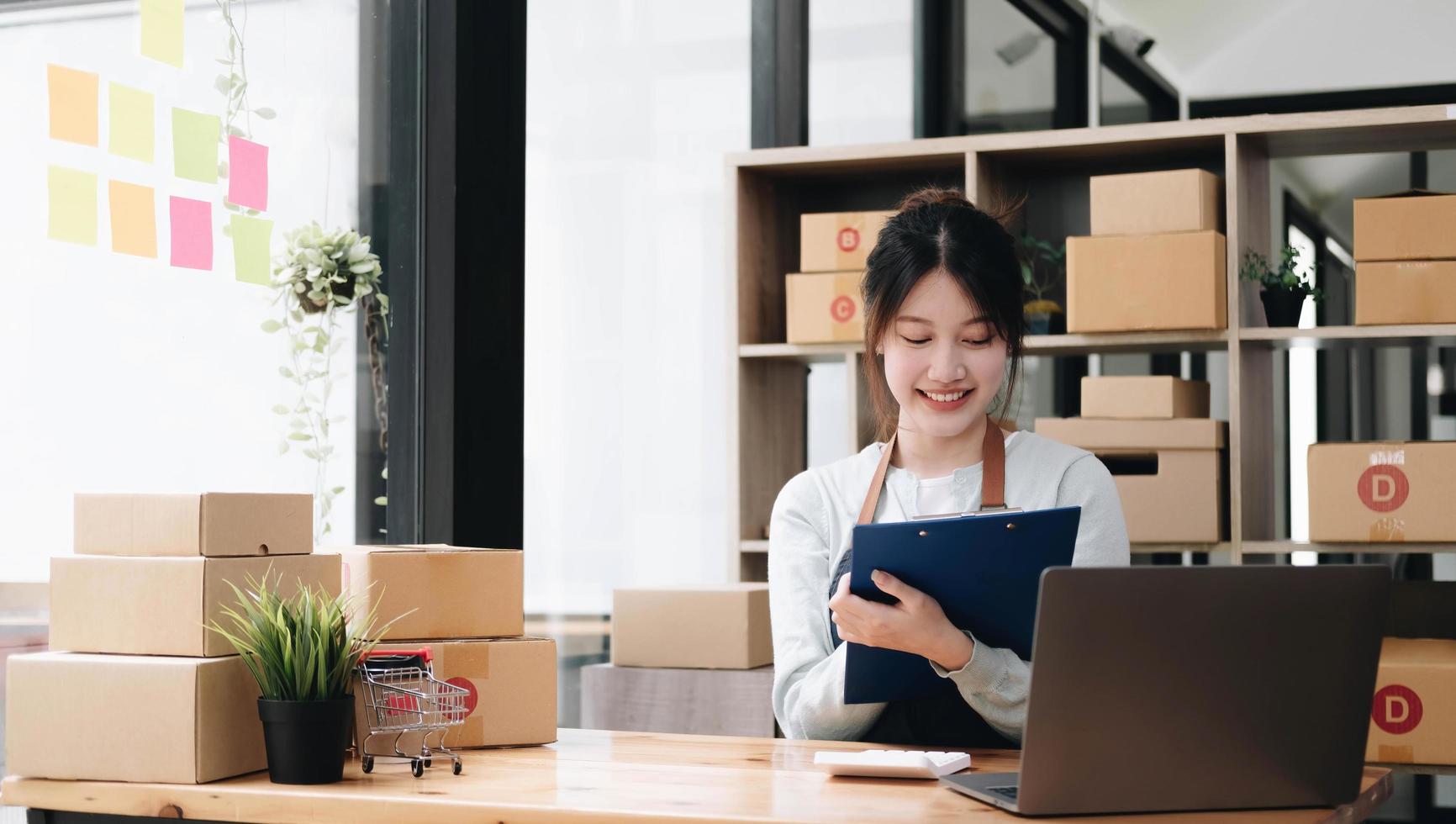 A portrait of a young Asian woman e-commerce employee sitting in the office full of packages in the background write note of orders and a calculator, for SME business e-commerce and delivery business. photo