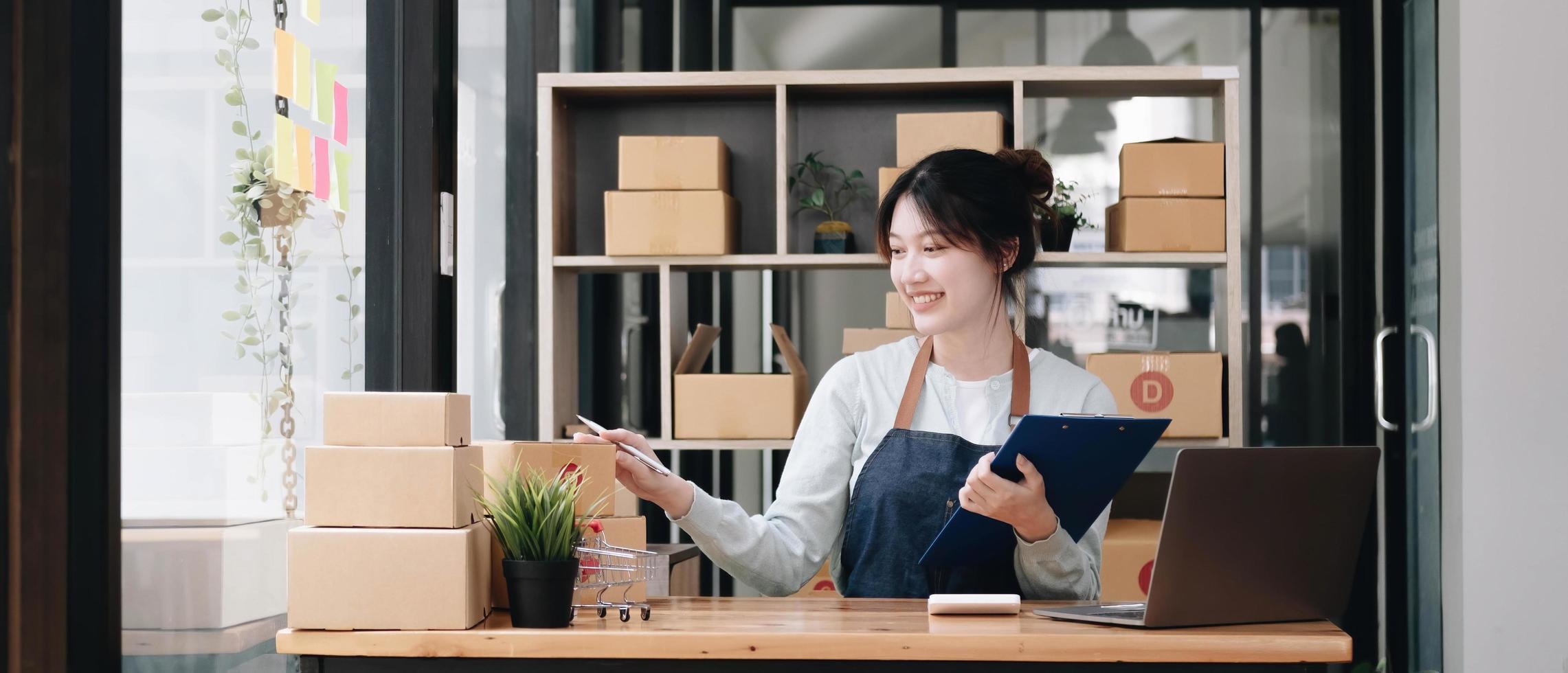 A portrait of a young Asian woman e-commerce employee sitting in the office full of packages in the background write note of orders and a calculator, for SME business e-commerce and delivery business. photo
