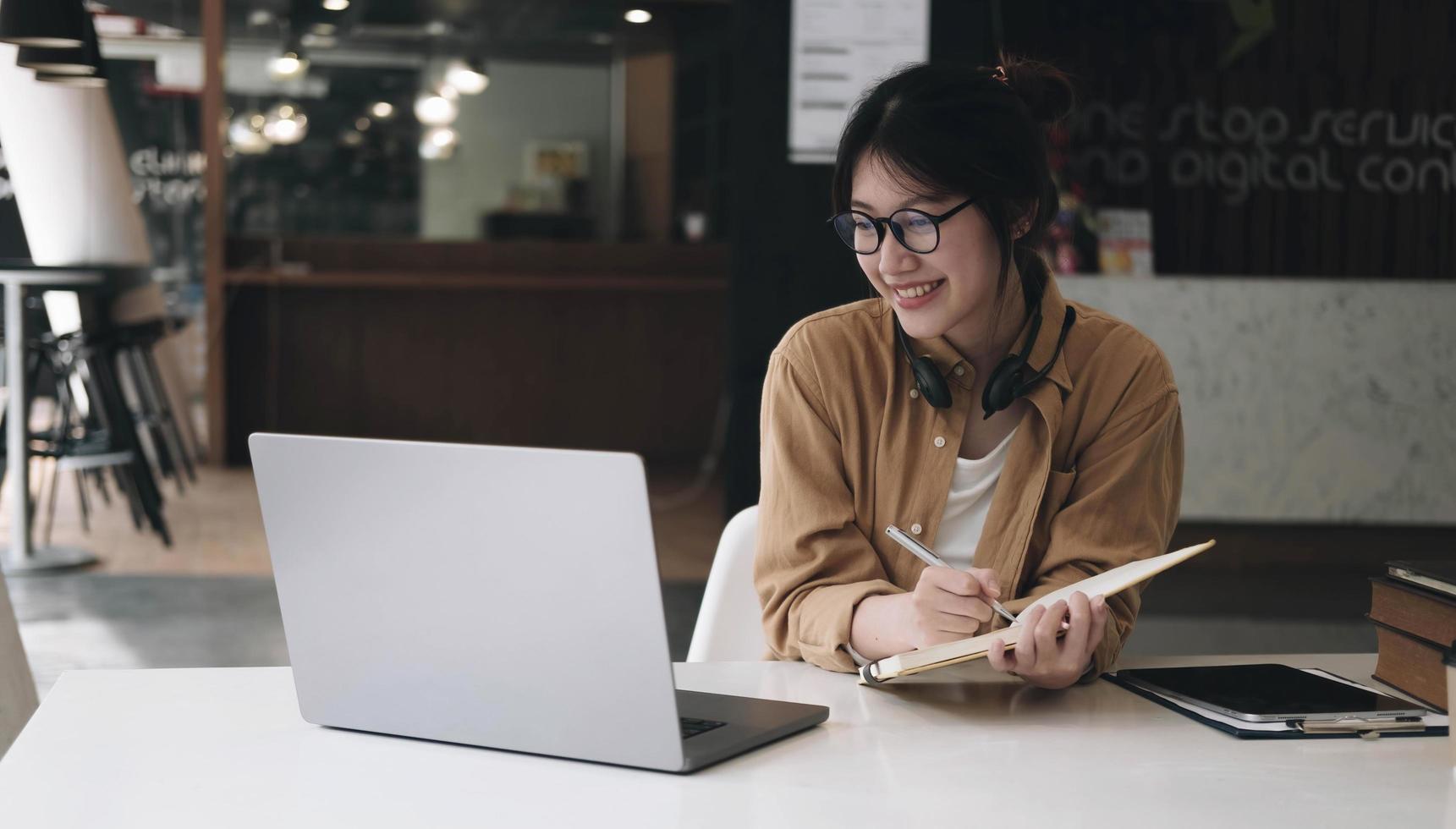 Happy asian businesswoman wearing headset make notes talk by conference call doing video chat on laptop in office, smiling chinese support service agent write information consult customer photo