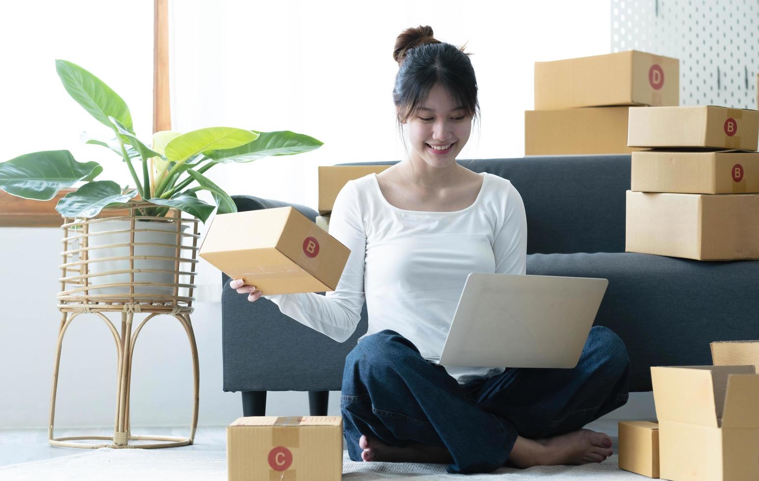 Smiling young Asian business owner woman prepare parcel box and standing check online orders of product for deliver to customer on laptop computer. Shopping Online concept. photo