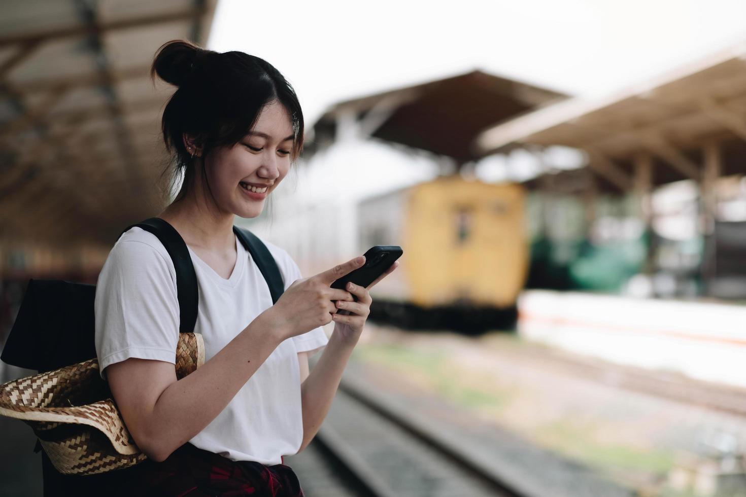 mujer esperando en la plataforma de la estación en el tren ligero de fondo usando un teléfono inteligente. mensaje de texto turístico y ruta del plan de parada de ferrocarril, transporte ferroviario, reservado. disfrutando del concepto de viaje foto