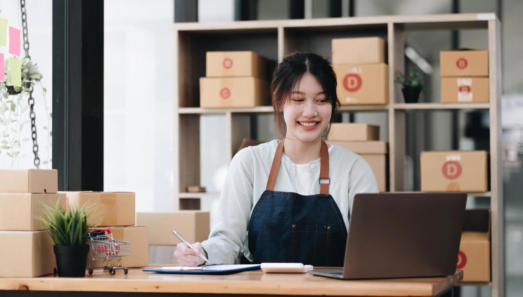 A portrait of a young Asian woman e-commerce employee sitting in the office full of packages in the background write note of orders and a calculator, for SME business e-commerce and delivery business. photo