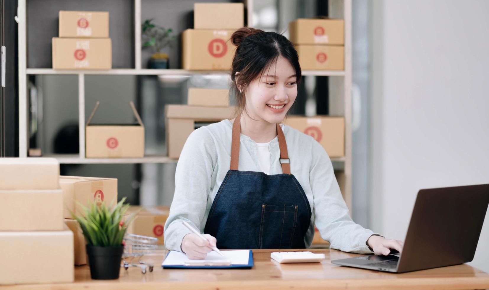 A portrait of a young Asian woman e-commerce employee sitting in the office full of packages in the background write note of orders and a calculator, for SME business e-commerce and delivery business. photo