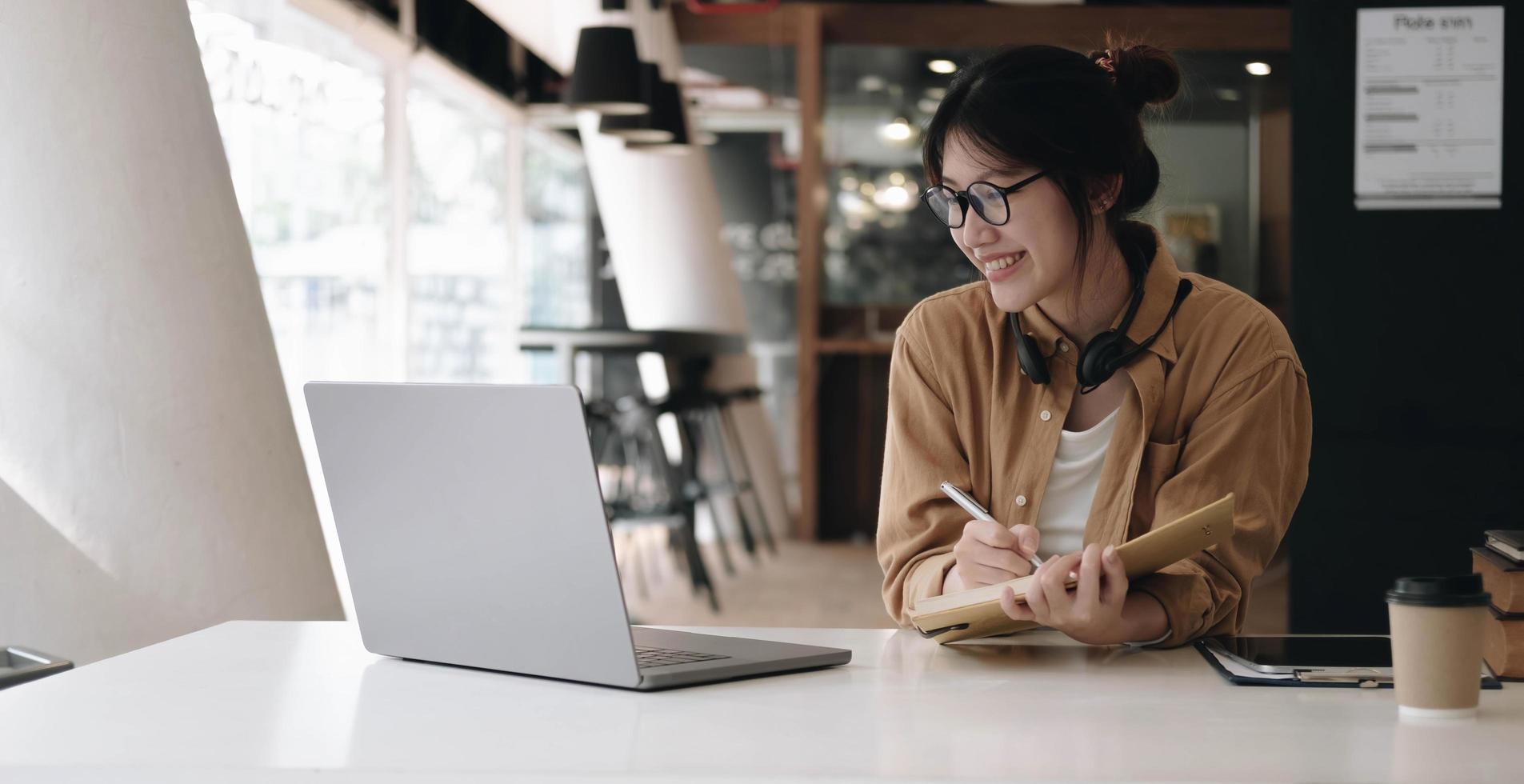 Happy asian businesswoman wearing headset make notes talk by conference call doing video chat on laptop in office, smiling chinese support service agent write information consult customer photo