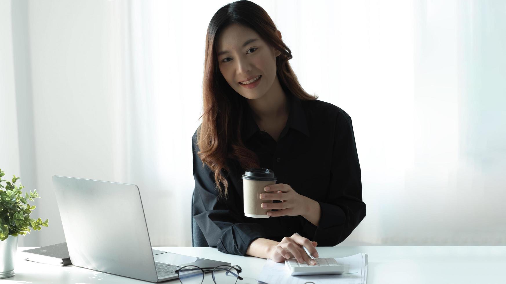Smiling Asian businesswoman holding a coffee mug and laptop at the office. Looking at the camera. photo