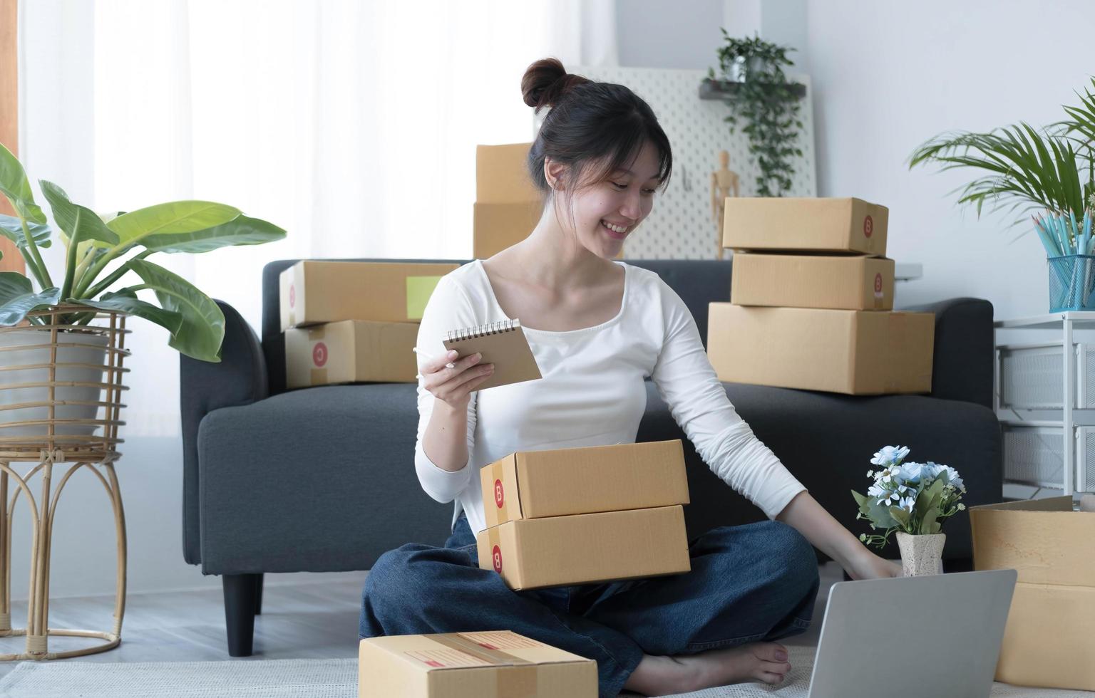 una joven y sonriente propietaria de una empresa asiática prepara una caja de paquetes y revisa de pie los pedidos de productos en línea para entregarlos al cliente en una computadora portátil. concepto de compras en línea. foto