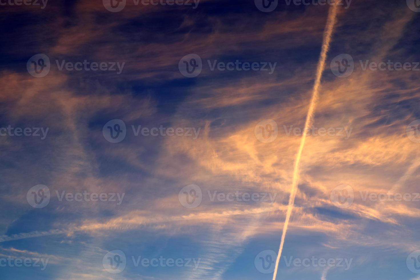 Aircraft condensation contrails in the blue sky inbetween some clouds photo