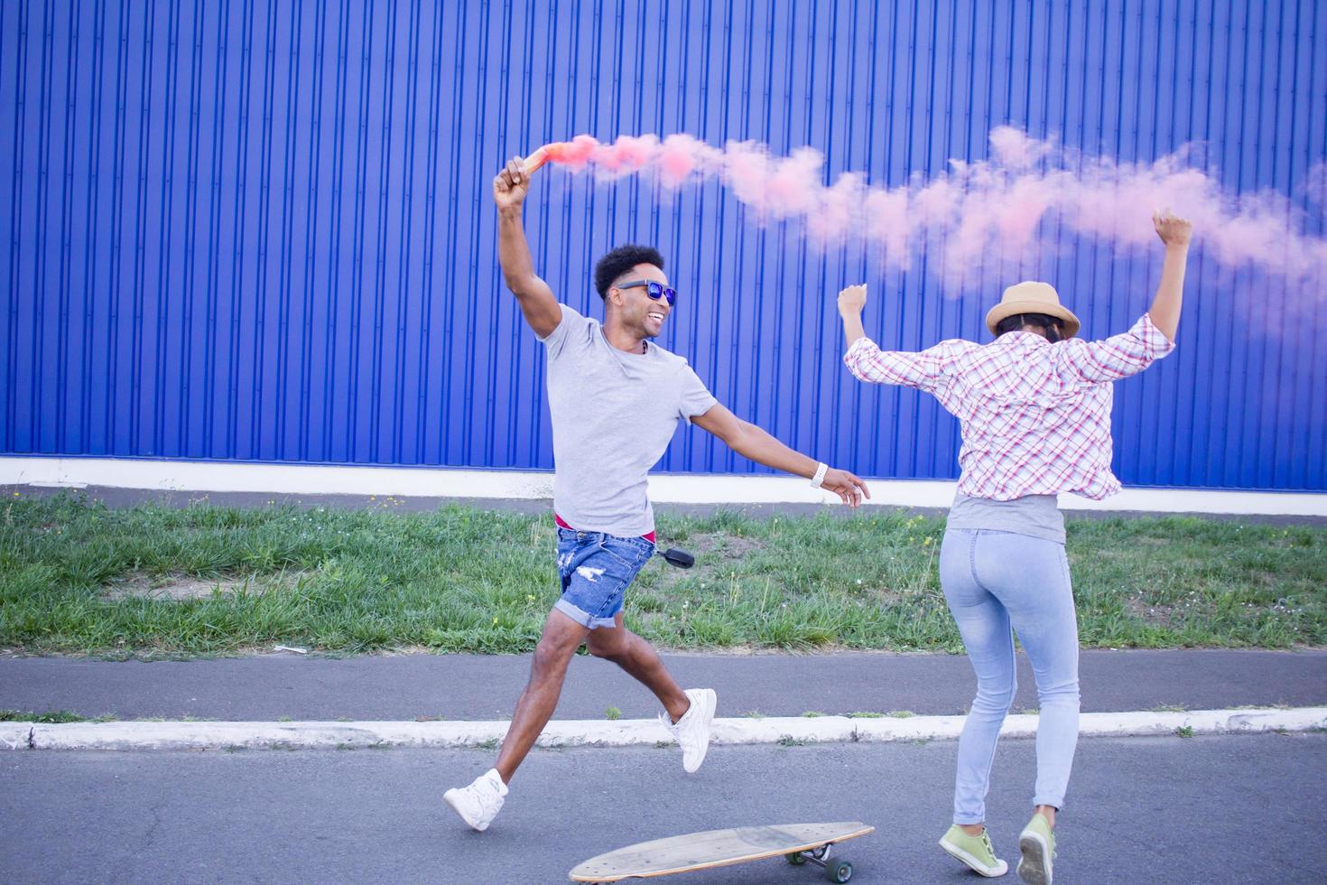 pareja joven montando en patinetas con bomba de humo de color, niño y niña con ropa informal pasando un buen rato, colores rosa y azul foto