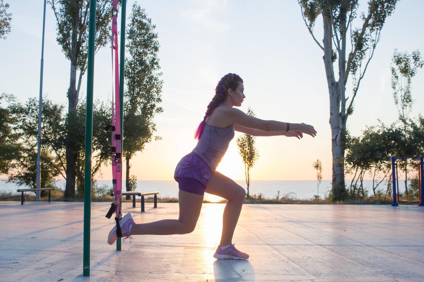 Beautiful fit woman in pink and purple sportwear training on outdoor gym in morning, exercises with suspension straps in park photo