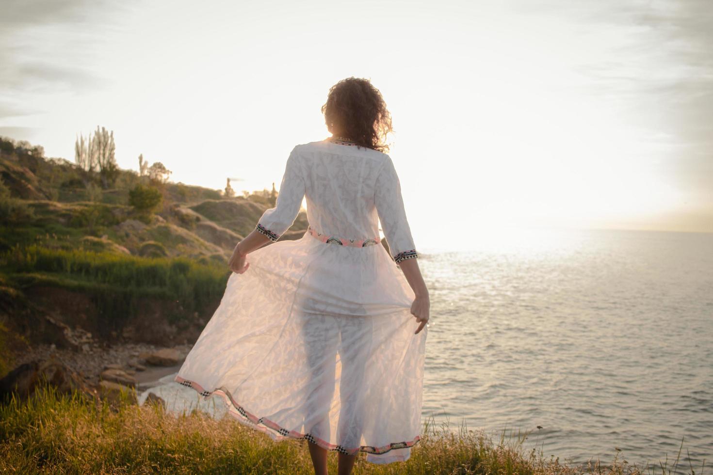 young woman walking on the morning beach in beautiful white dress. Fit female having good time during turing the sunrise. photo