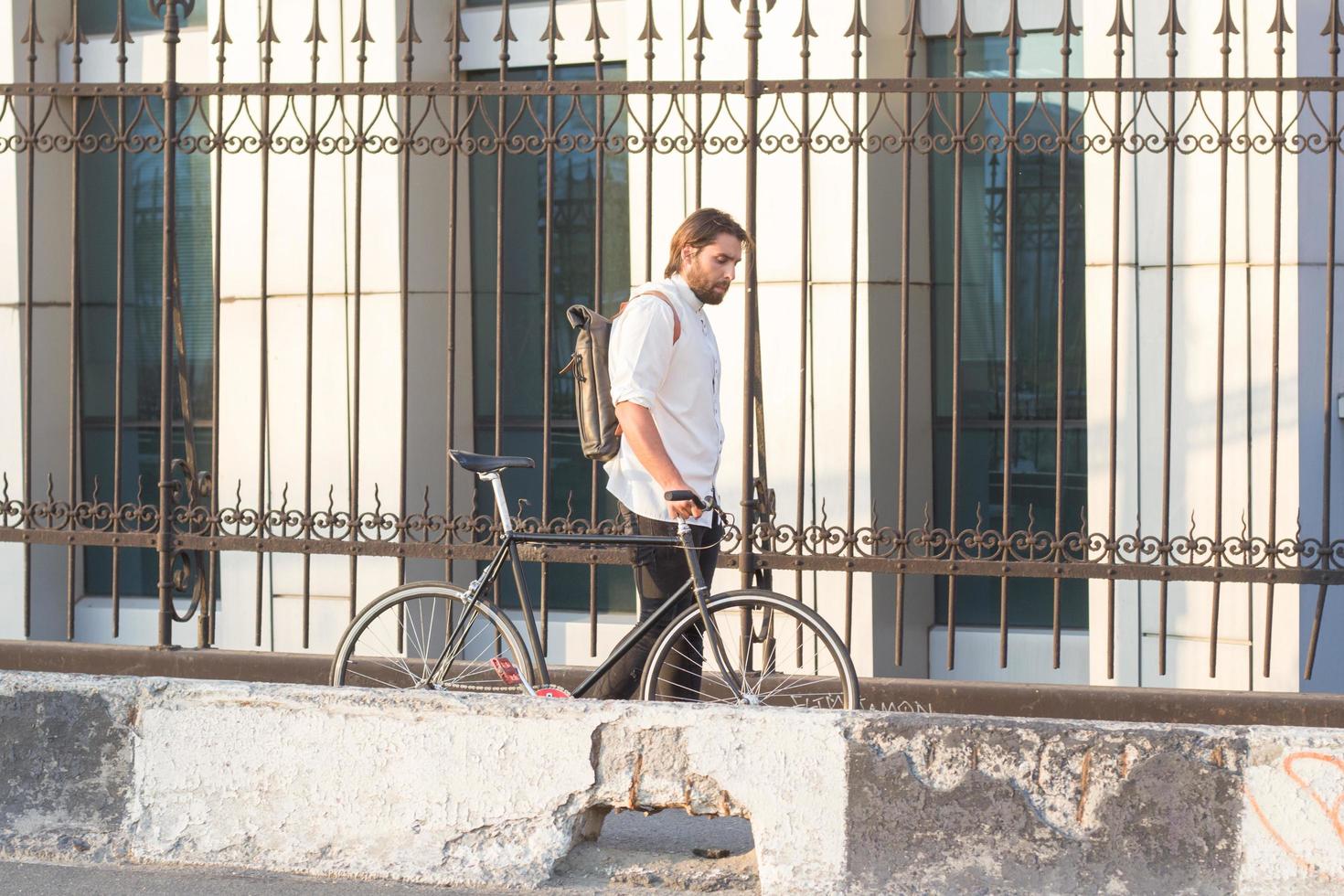 Young bearded man with leather backpack walking with black bicycle photo