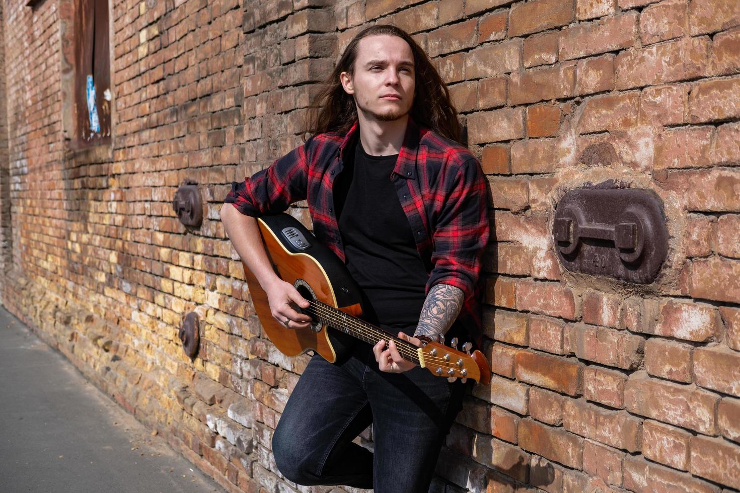 Young male with long hair and tattoo play on acoustic guitar outdoors on the street photo