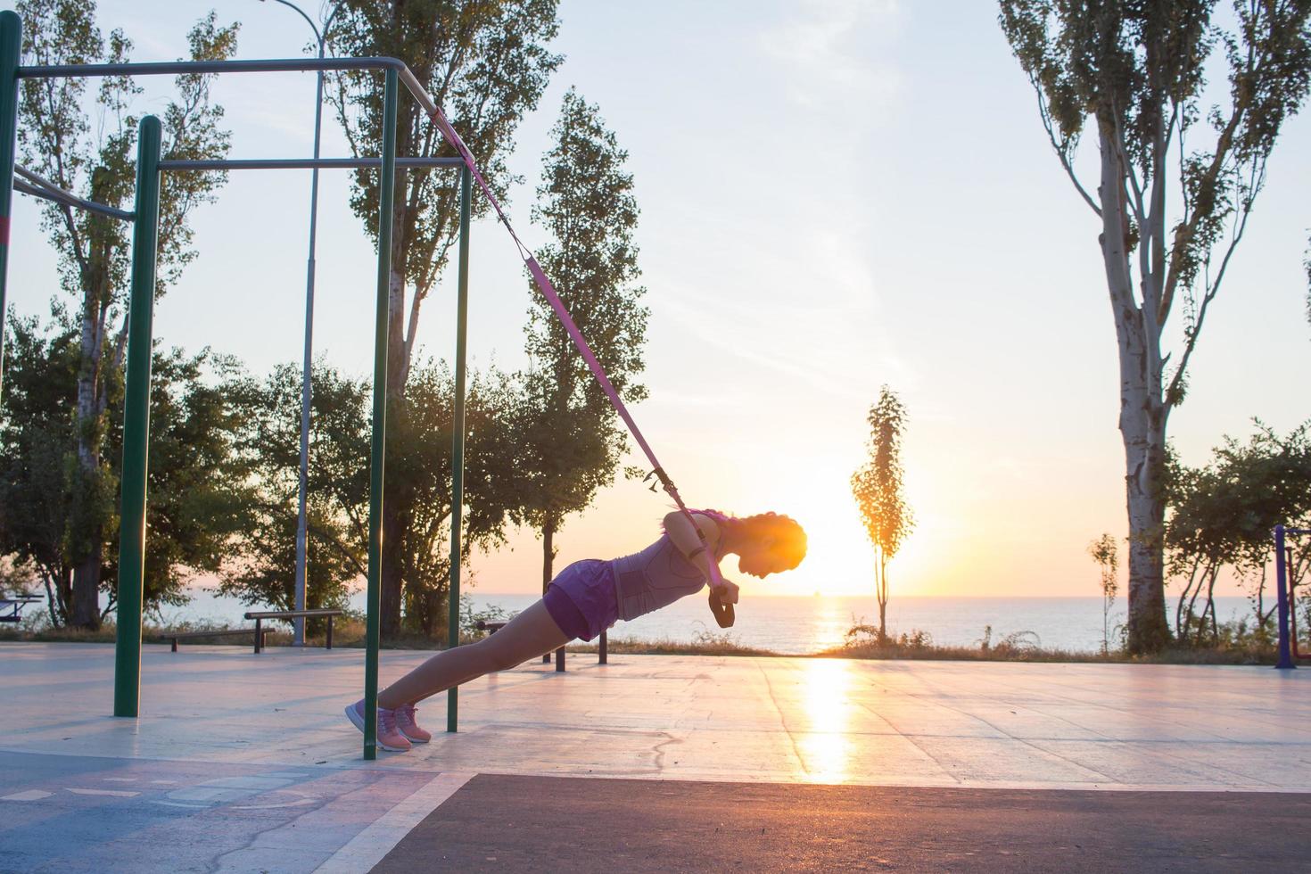 hermosa mujer en forma en ropa deportiva rosa y púrpura entrenando en el gimnasio al aire libre por la mañana, ejercicios con correas de suspensión en el parque foto