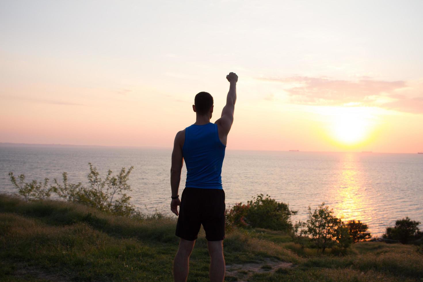 Happy fit runner watching sunrise or sunset with rised fists, young athlete on grass during the sunrise in the sea photo