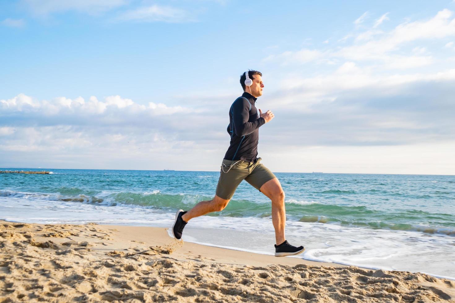 ajuste el entrenamiento de corredores masculinos en la playa de verano y escuche música contra el hermoso cielo y el mar foto