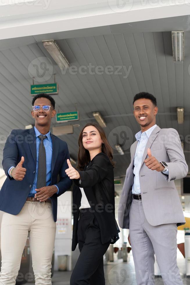 Group of business people thumbs up for acceptance. Confident young business people in full suit standing outdoors looking away with cityscape photo