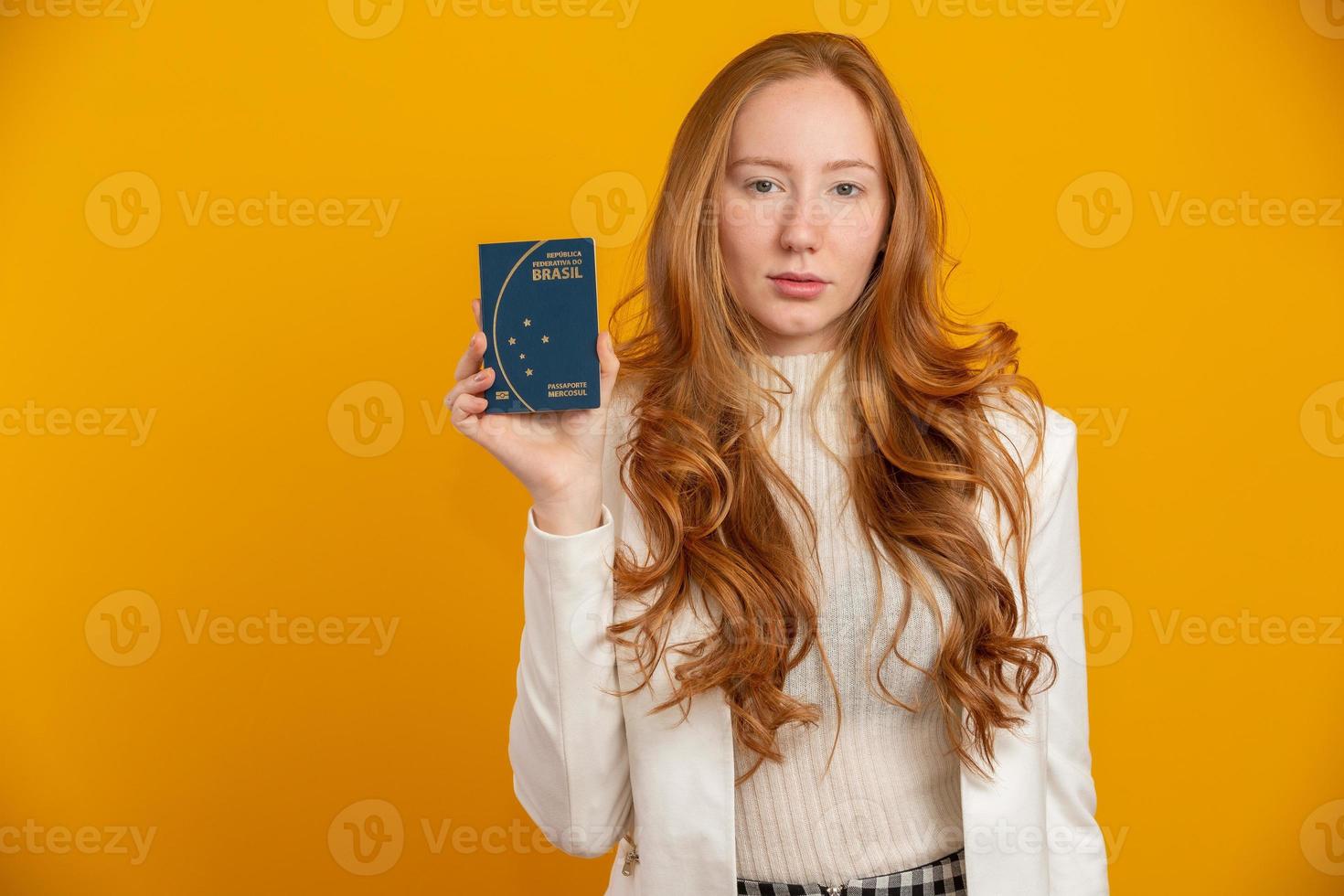 Beautiful redhead curly-haired girl in travel. Next trip. Girl holding Brazilian passport. On yellow. photo