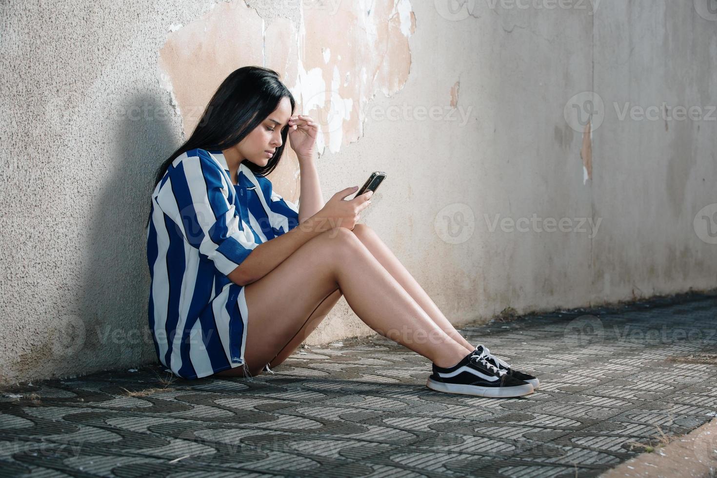 Single sad teen holding a mobile phone lamenting sitting on the sidewalk. Crying depressed teen girl holds phone sitting on the sidewalk. photo