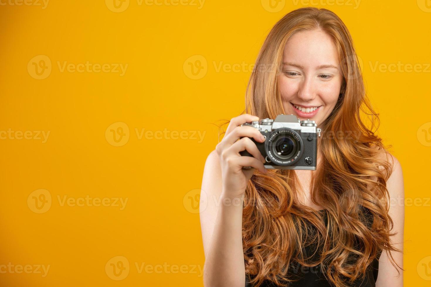 Photo of adorable redhead woman wearing black clothes holding retro vintage camera and taking picture isolated over yellow background. Mock up copy space. Hold retro vintage photo camera.