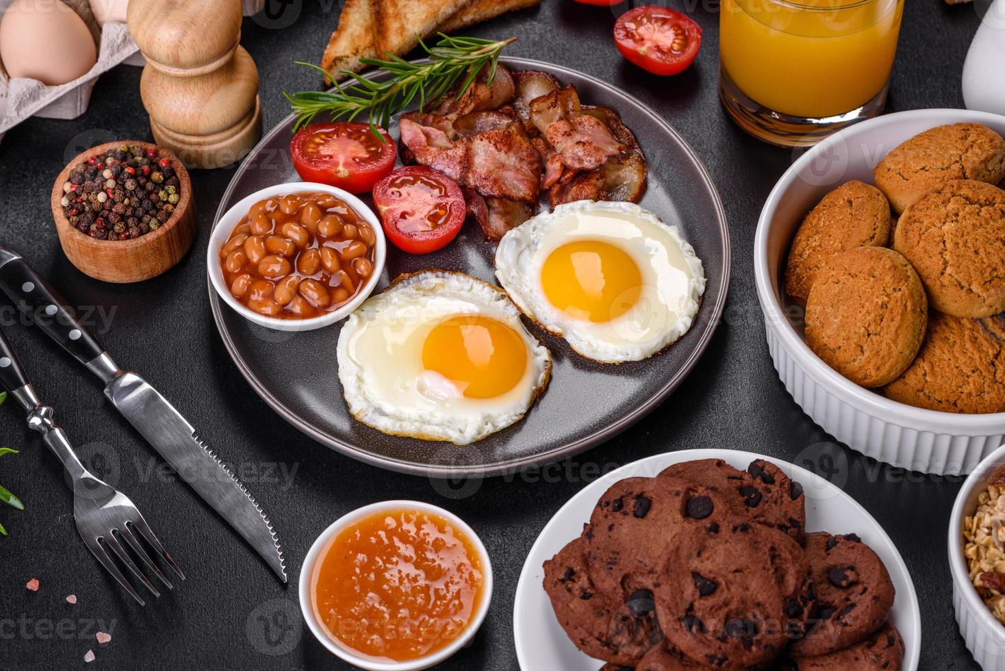 un delicioso desayuno saludable y nutritivo con huevos fritos, tocino, frijoles, un vaso de jugo, galletas de avena, leche y mermelada foto