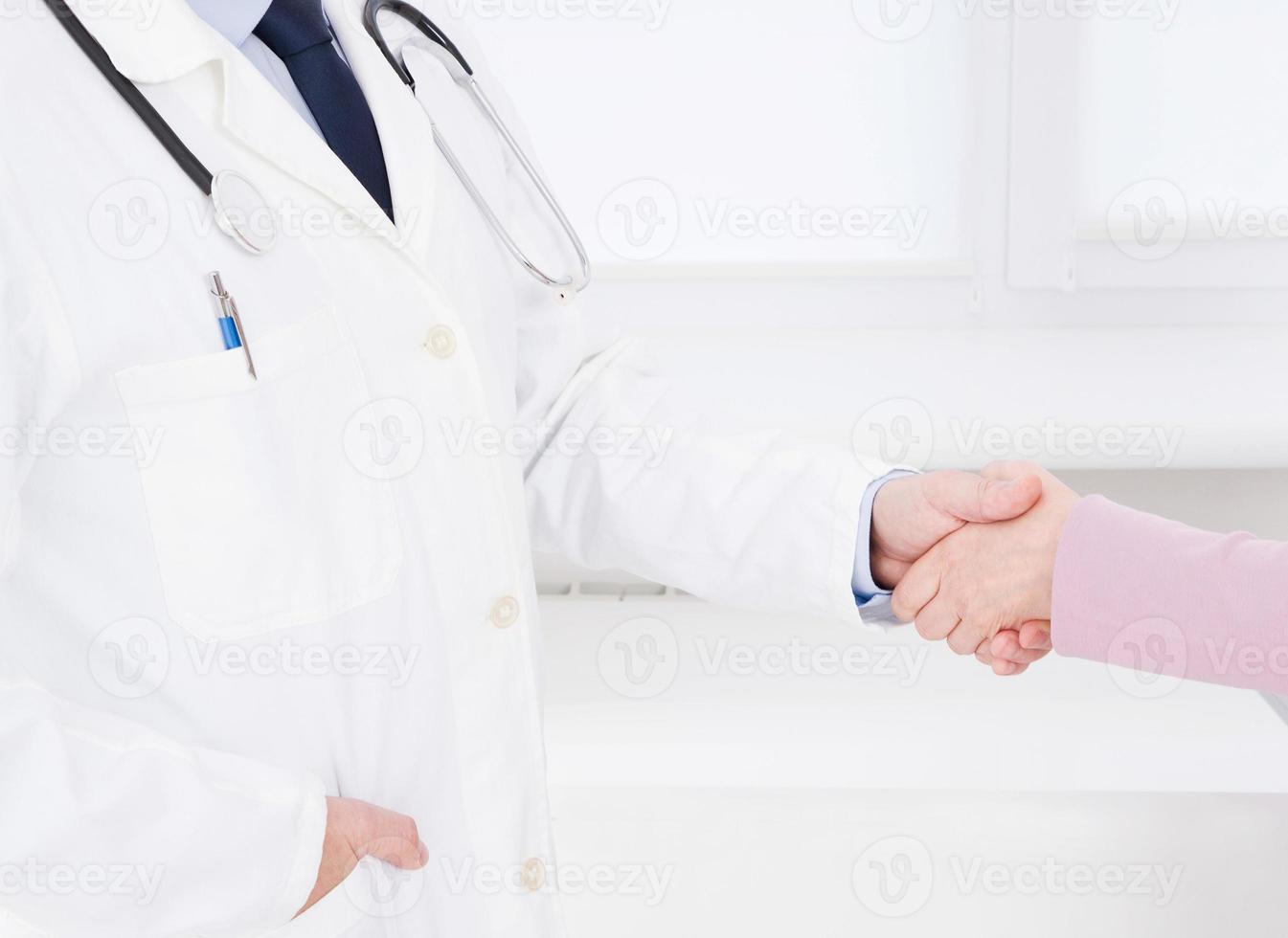 Doctor and patient in the office shaking hands, healthcare and assistance concept.Medical insurance. Doc sitting in cabinet. Man in white uniform. photo