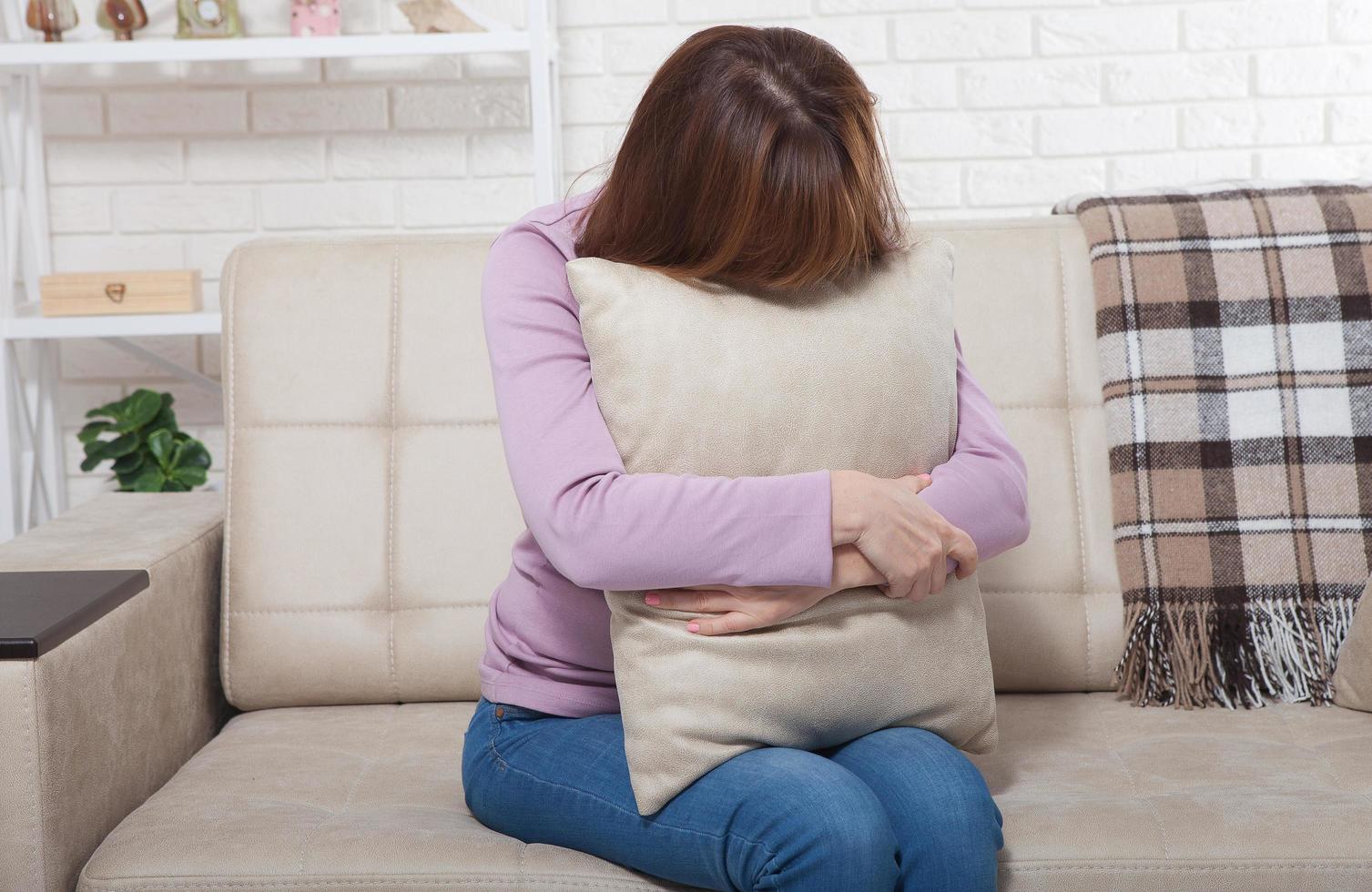 Beautiful middle aged brunette woman holding a pillow and crying on sofa. Home background. Menopause time photo