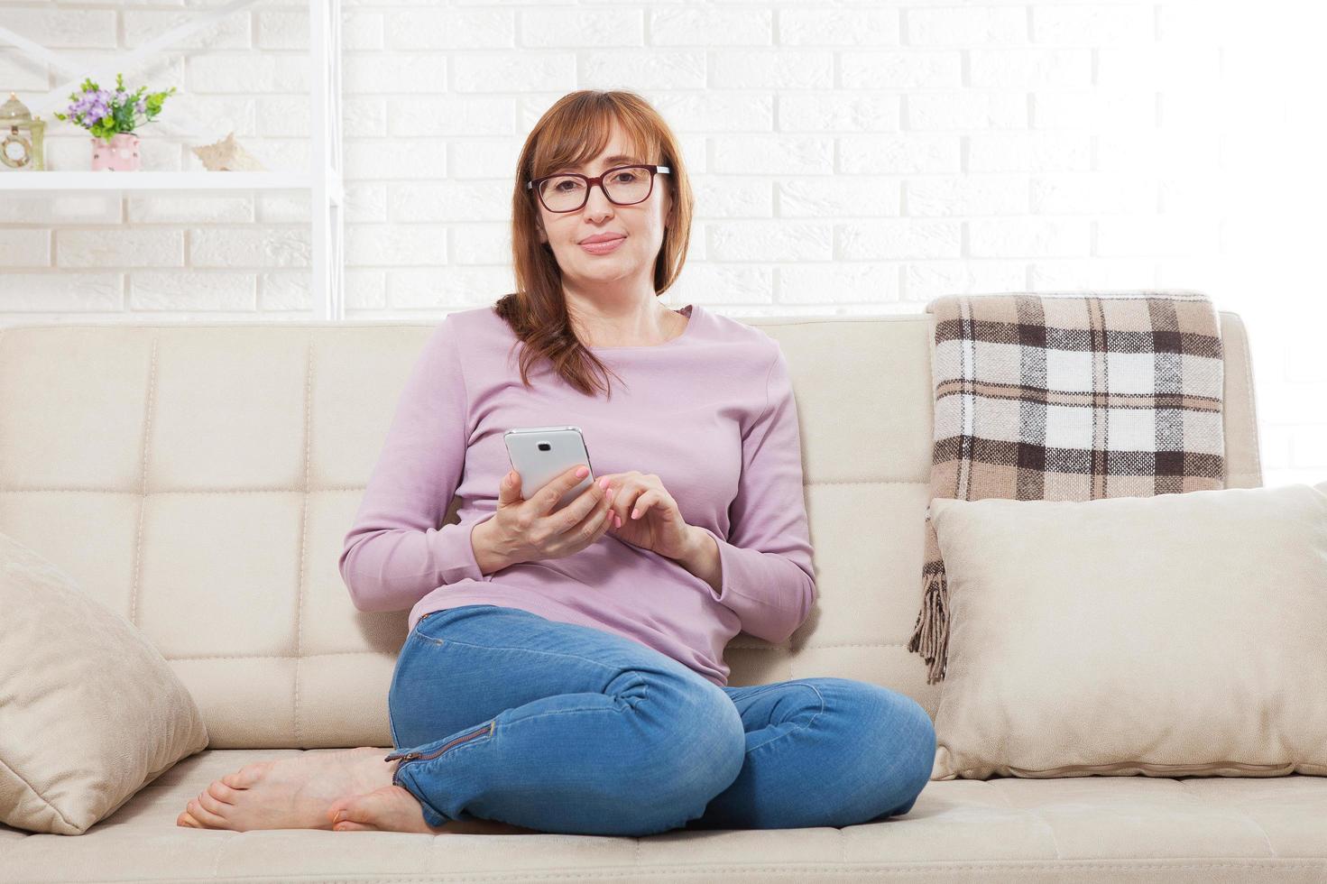 Smiling middle age Woman sitting on sofa and typing message on smart phone at home. Housewife using online shopping on mobile phone. Mock up and copy space photo