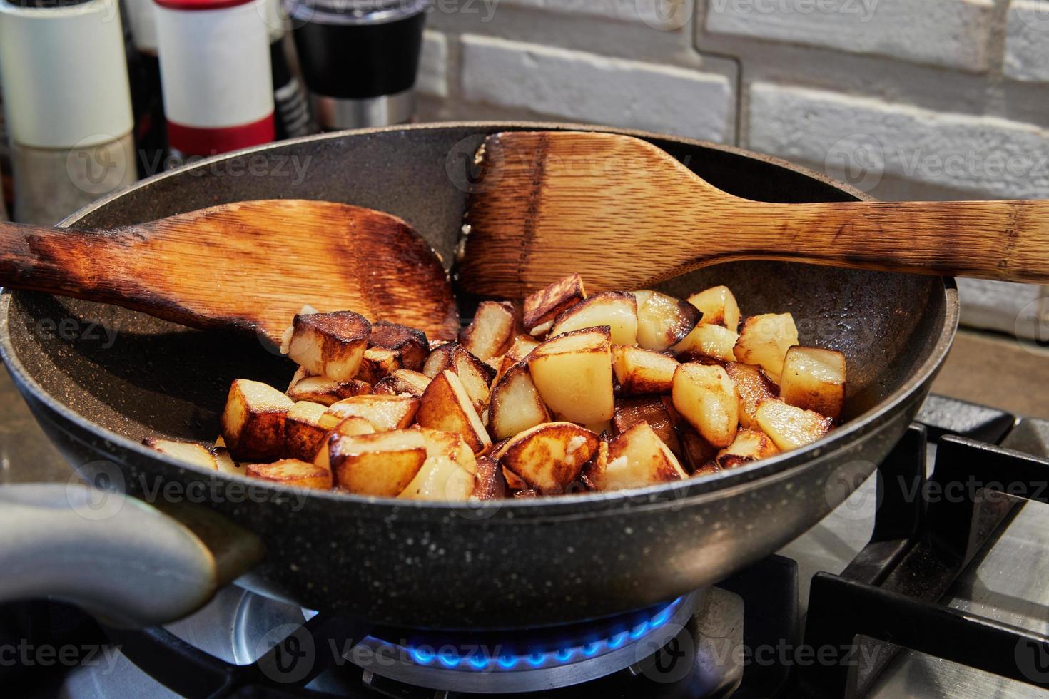 Frying pan with fried potatoes and wooden spatulas on gas stove. Step by step recipe photo