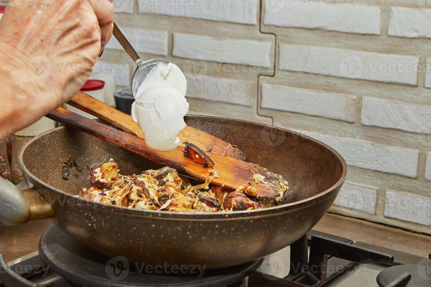Chef pours fresh cream into pan with fried potatoes, chestnuts, parsley, parmesan and wooden spatulas on gas stove. Step by step recipe photo