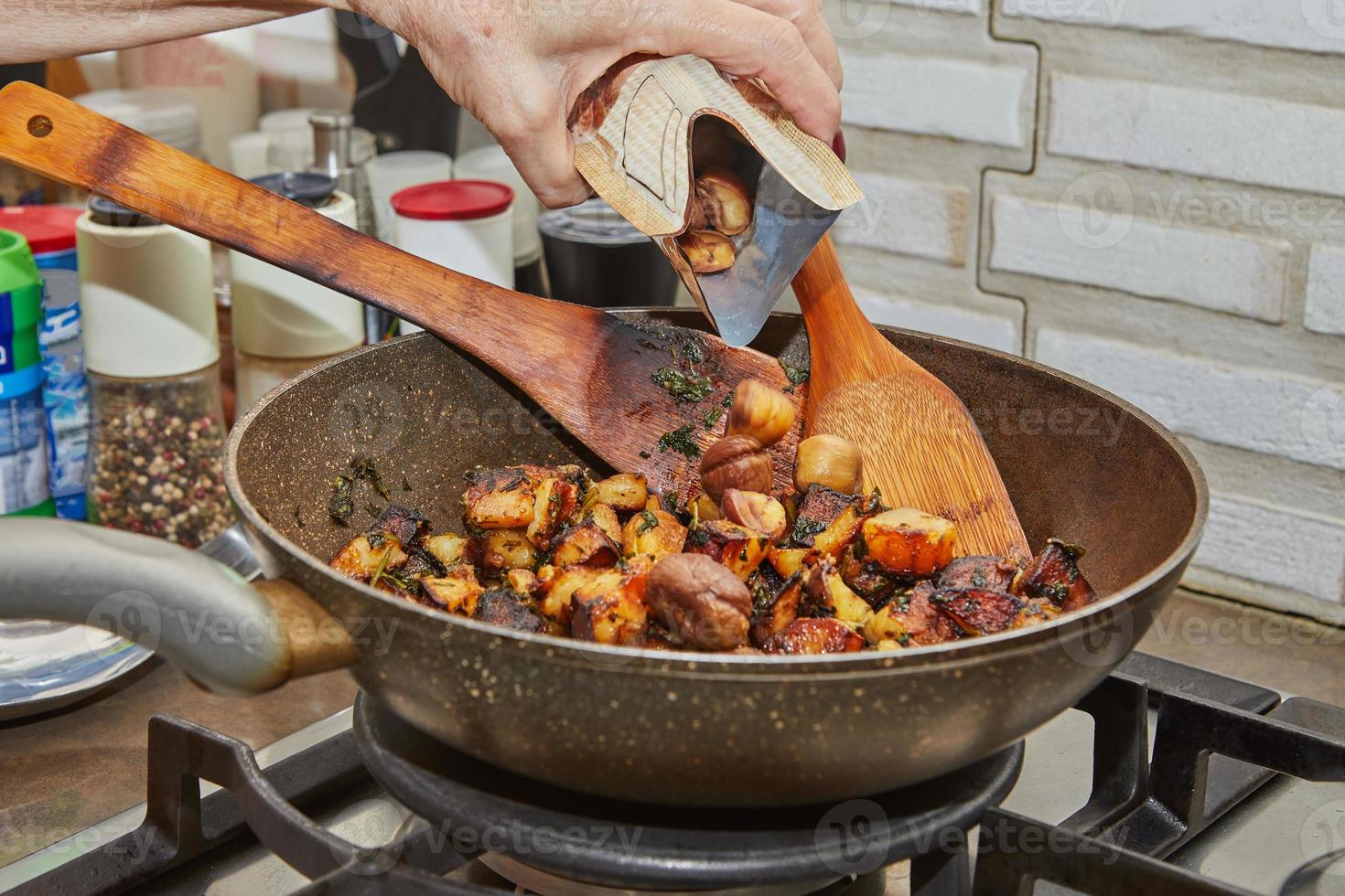 el chef vierte castañas en una sartén con papas fritas, perejil y espátulas de madera en la estufa de gas. receta paso a paso foto