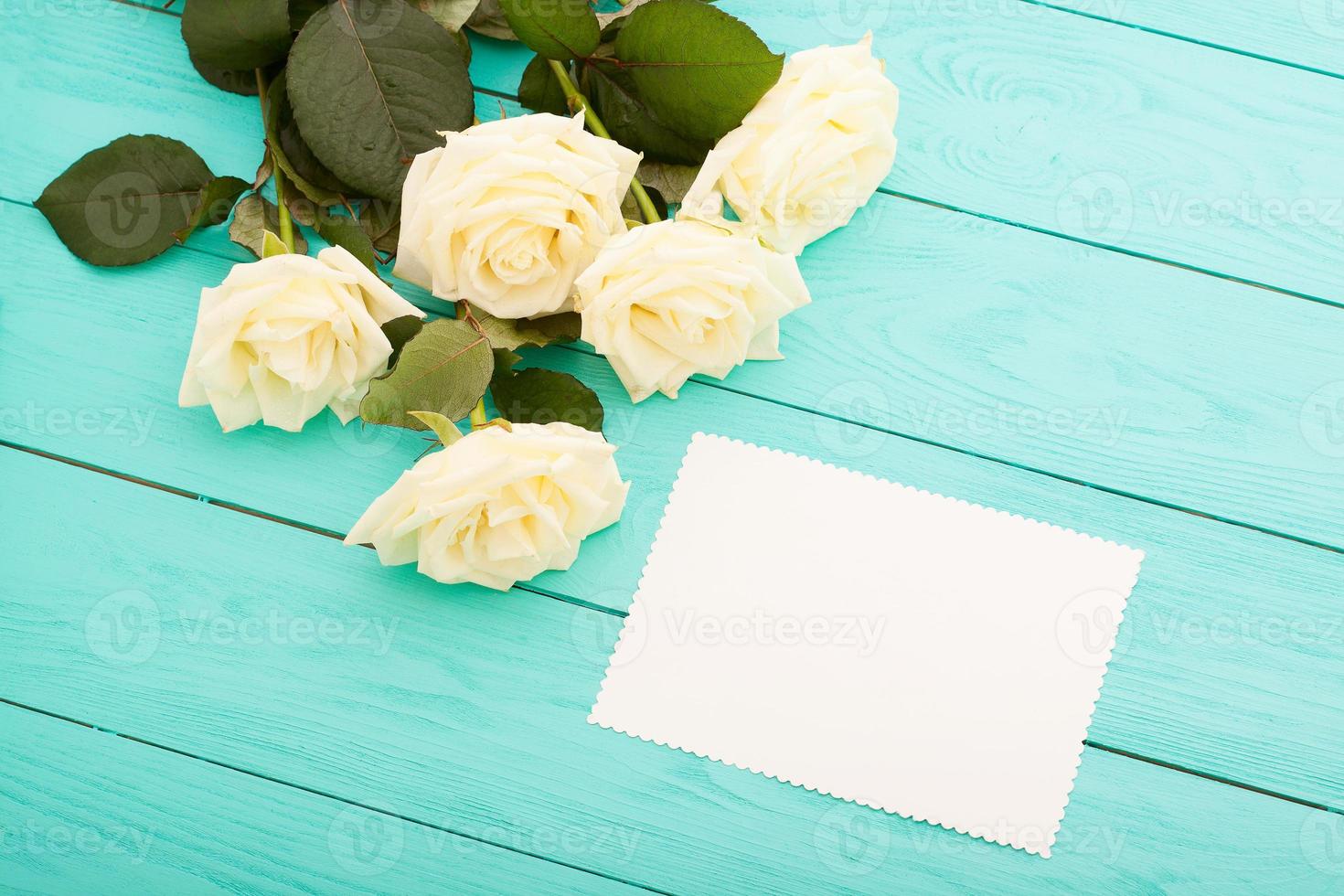 Frame of colorful roses on blue wooden background with paper card and copy space. Top view and selective focus. Mock up photo