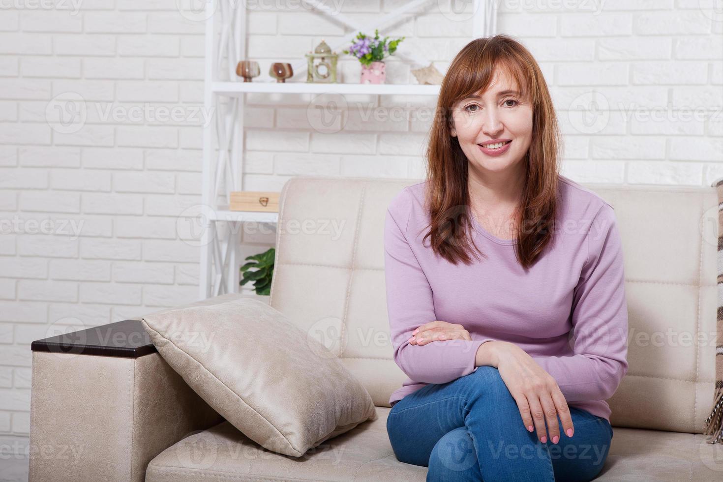 Beautiful and cute smiling middle age woman sitting on sofa. Home background. Happy menopause concept. photo