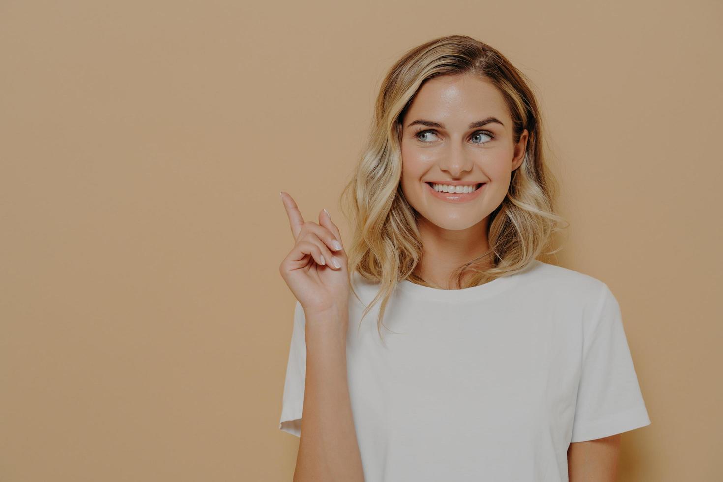 cliente joven positiva con cabello rubio usando camiseta blanca con una sonrisa agradable apuntando al espacio de copia foto