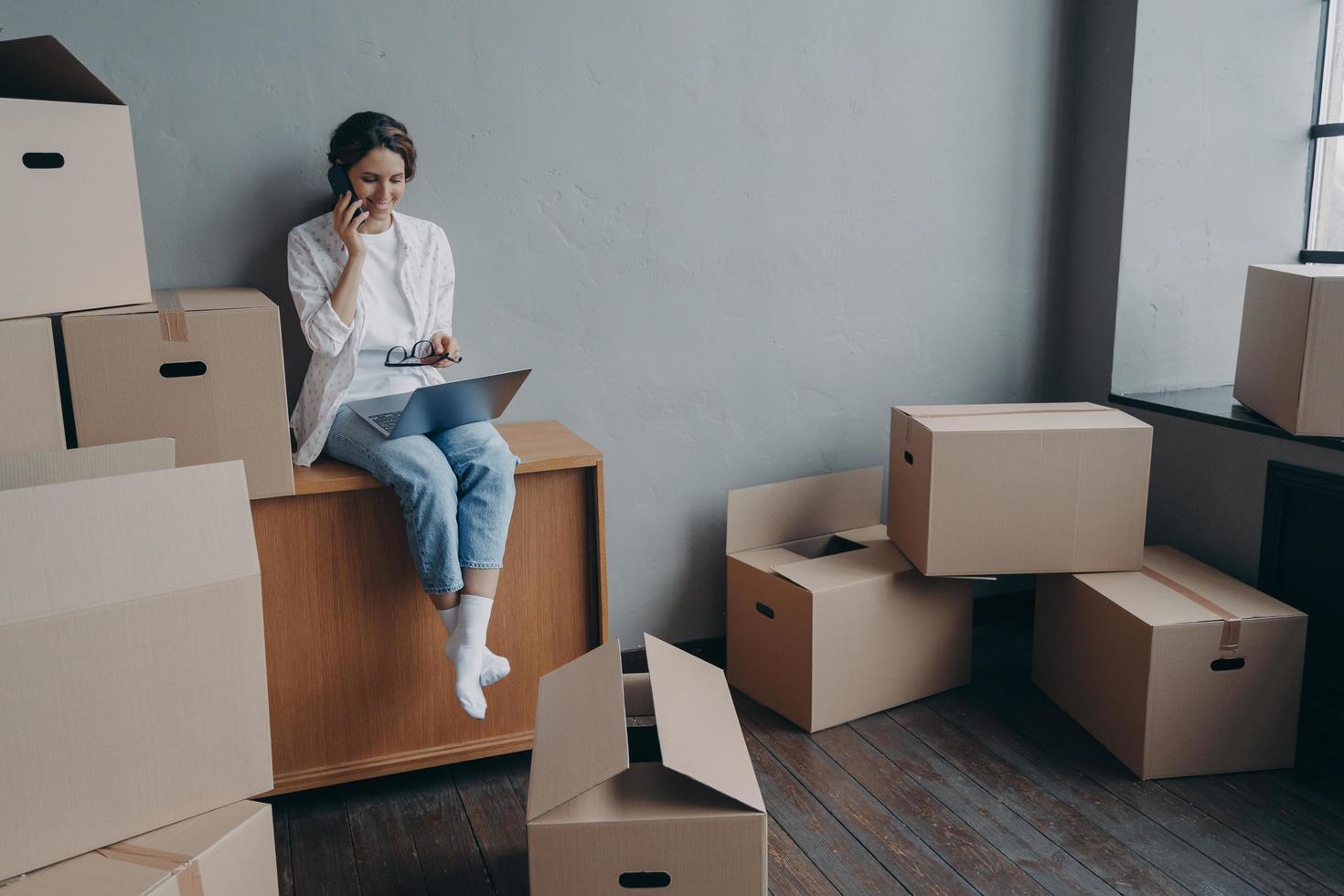 Happy homeowner talking on phone. Hispanic girl is sitting among carton boxes and working on pc. photo