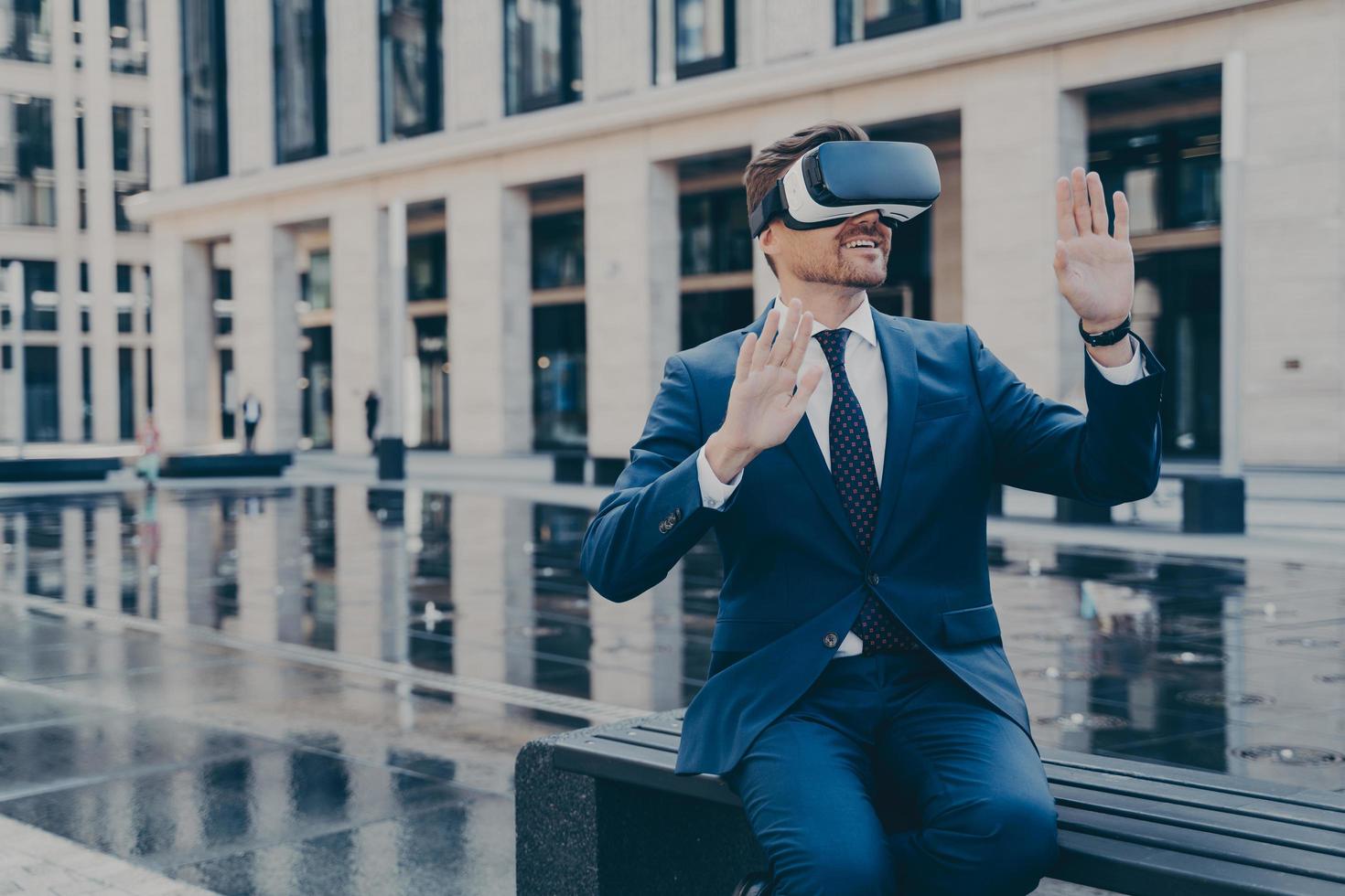 Man in VR goggles trying to reach something in virtual reality with his hands while sitting outdoors photo
