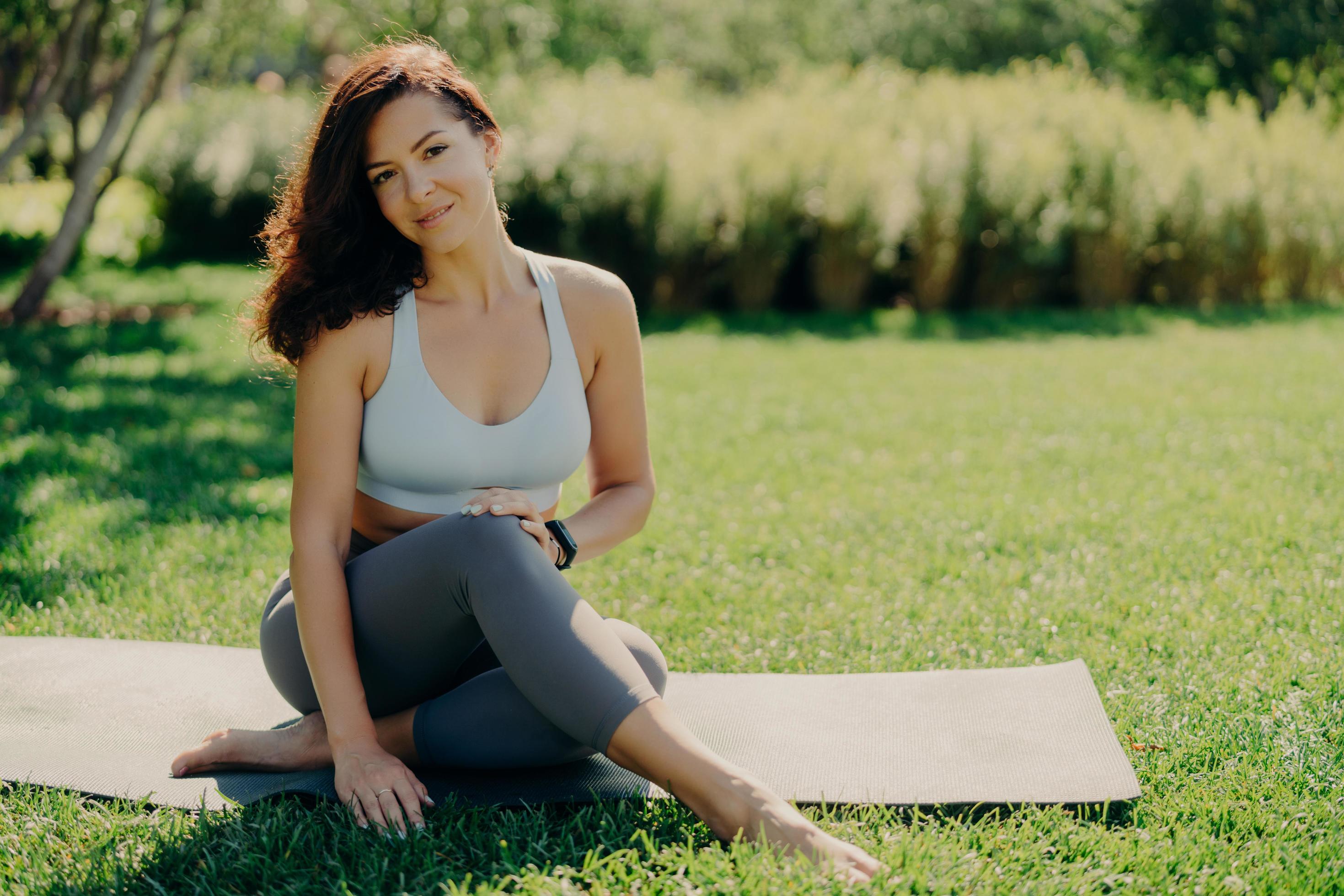 Photo of brunette sportswoman has rest after fitness training sits on  karemat with bare feet in activewear leads healthy lifestyle poses on fresh  green grass enjoys summy day and good weather 7662092