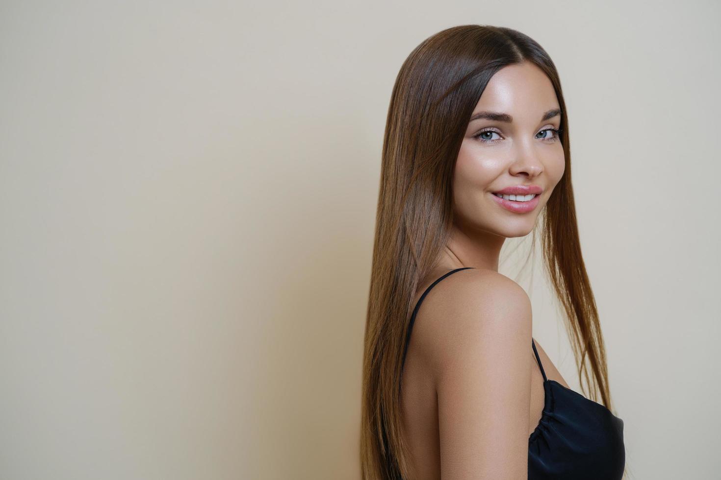 Portrait of beautiful tender girl with well cared long dark hair, has bare shoulder, dressed in black dress, has pleased expression, isolated over beige background. Beauty, makeup, personal care photo