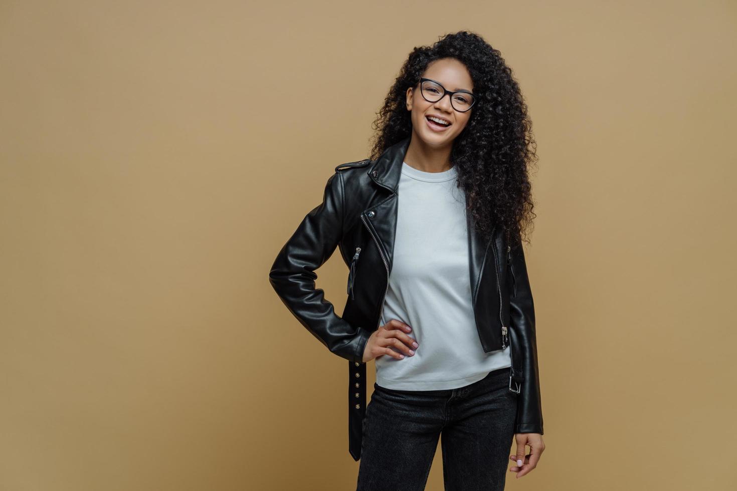Fashionable woman with glad expression, keeps hand on hip, smiles cheerfully, wears optical glasses, white t shirt, black leather jacket and jeans, ready for walk, poses against beige background photo