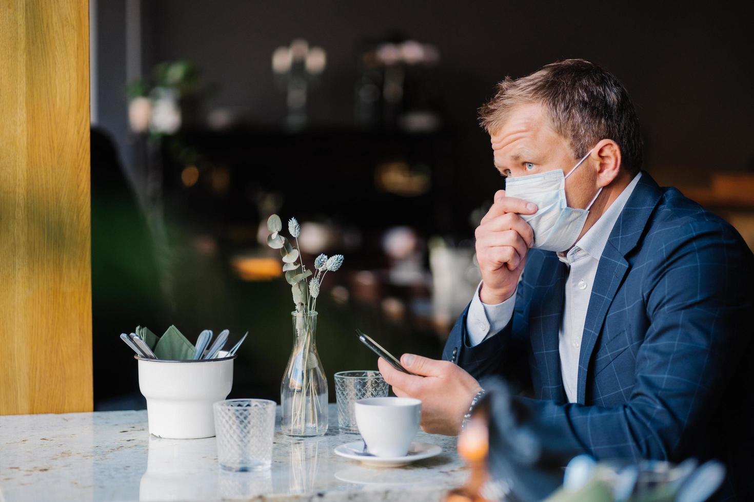 Elegant businessman coughs and wears protective medical mask during epidemic situation, sits in cozy cafeteria, waits for call, holds modern cell phone. Coronavirus pandemic, health care concept photo