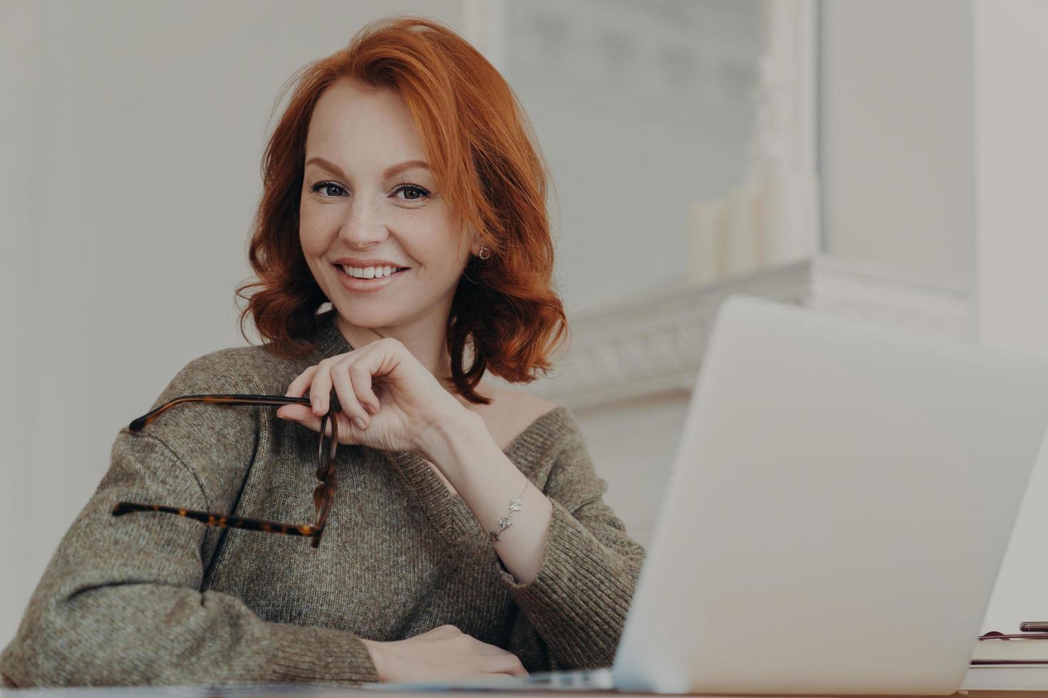 Skilled successful redhead woman holds eyewear for vision protection, sits in front of opened laptop computer, poses at coworking space, wears sweater, uses modern device. Distance work concept photo