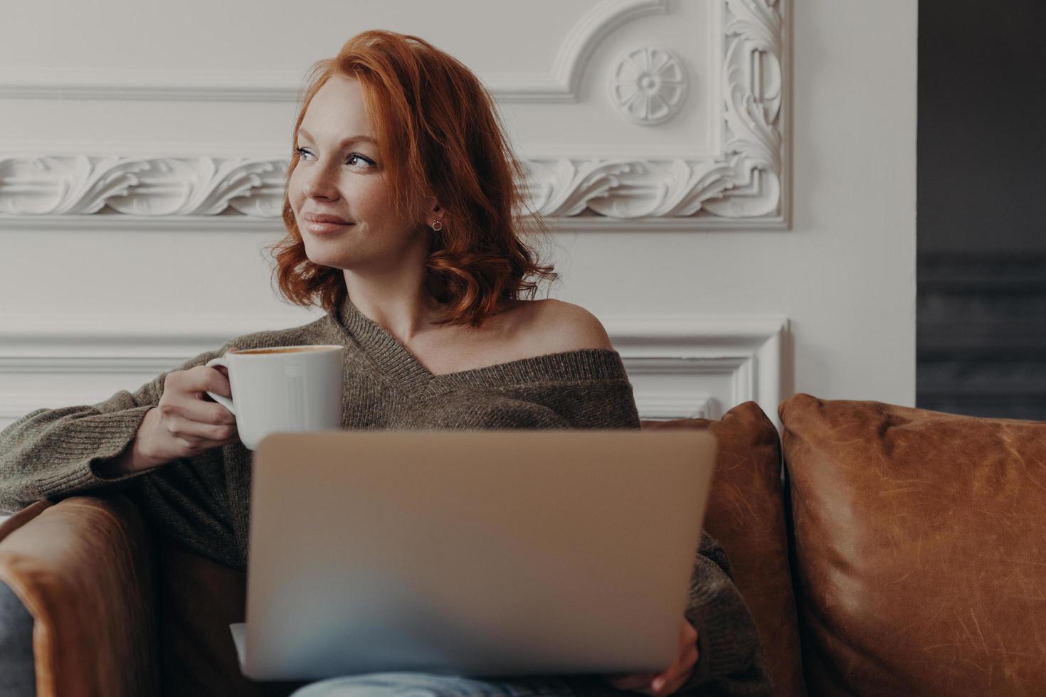 Indoor shot of satisfied redhead woman works freelance on laptop computer, drinks aromatic beverage, concentrated aside with thoughtful expression, sits on comfortable sofa, thinks about new project photo