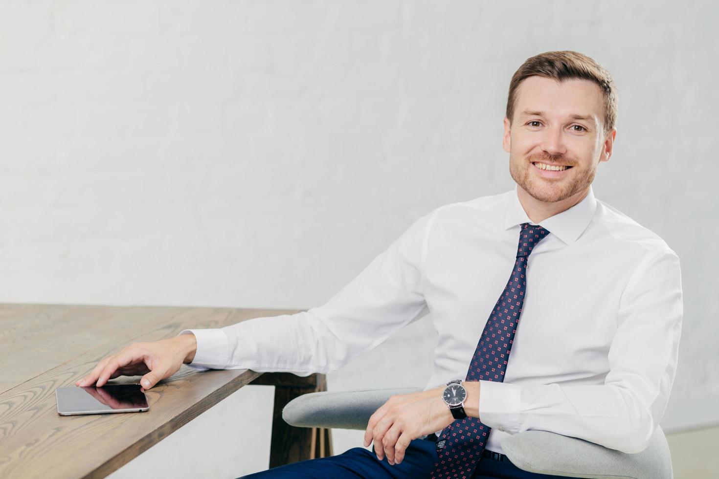 foto de un joven empresario positivo y exitoso vestido con un elegante pantalones blanco con corbata, mira positivamente a la cámara, tiene un descanso después del trabajo, posa en su gabinete, usa una tableta moderna