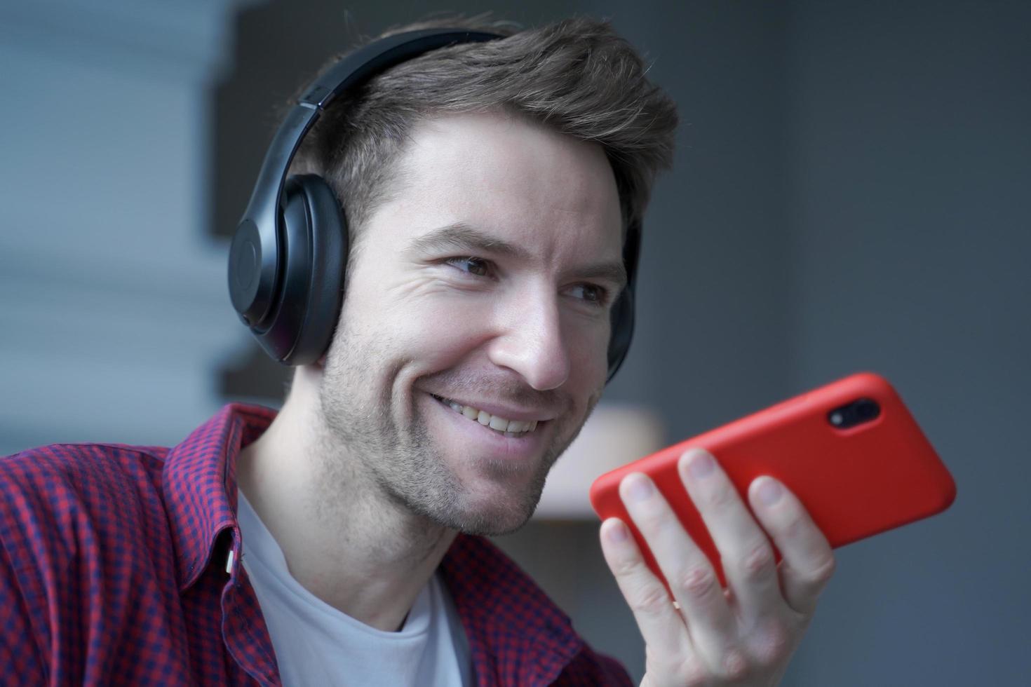 Cheerful young german man in wireless headphones holding smartphone in hand, recording audio message photo